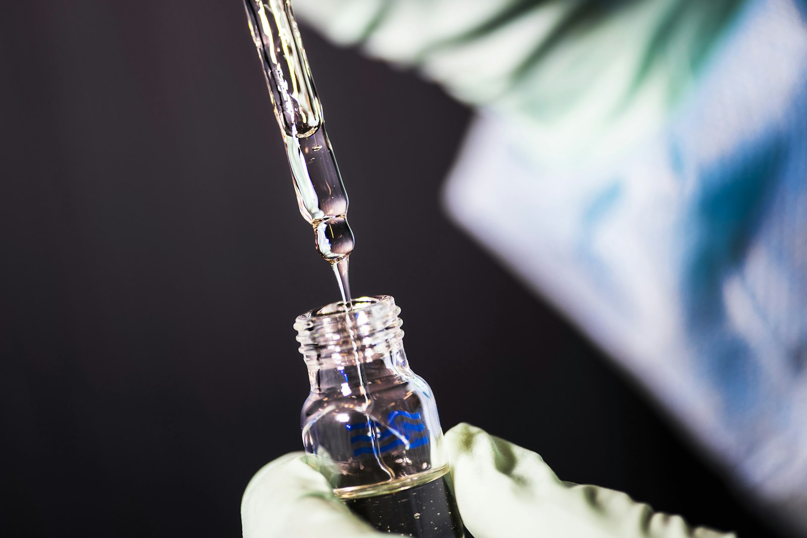 Gloved hand using a pipette to dispense liquid into a small sample vial.
